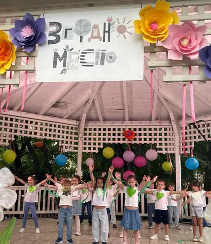Children on stage with arms raised, wearing green paper leaves and ribbons