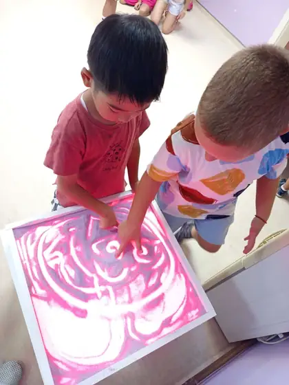 Two boys drawing patterns in pink sand on a light table