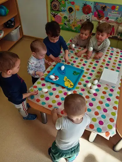 Toddlers gathered around a low table, dyeing white eggs on sticks