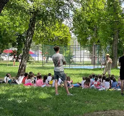 Children sitting on the grass in a park while a young man leads an outdoor game