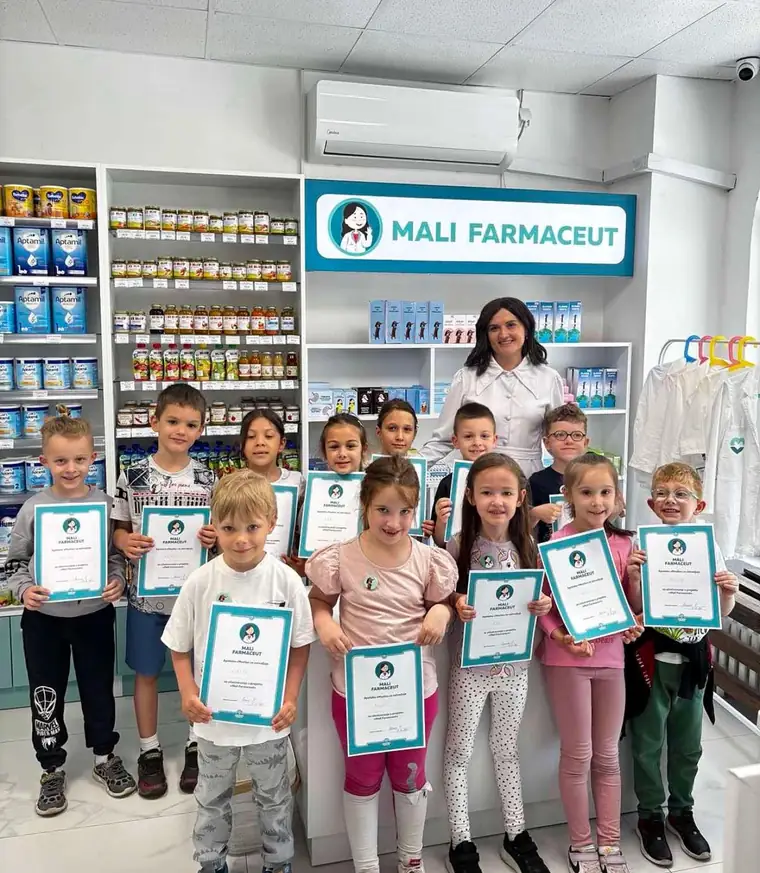 Deca drže diplome „Mali farmaceut“ ispred polica sa lekovima, uz farmaceutkinju u belom mantilu